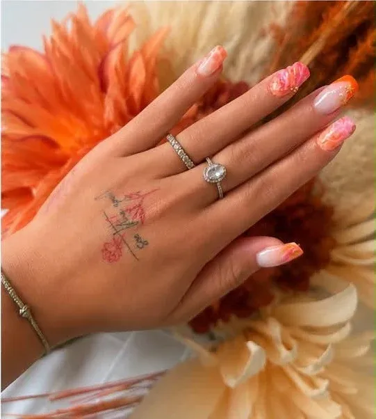 Main aux ongles effet aquarelle corail, bagues et fleurs orangées en fond